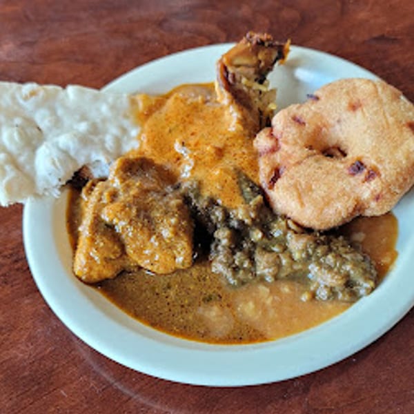 A white plate of Daddy's Indian Cuisine with chicken curry, a fried vada, and flatbread.
