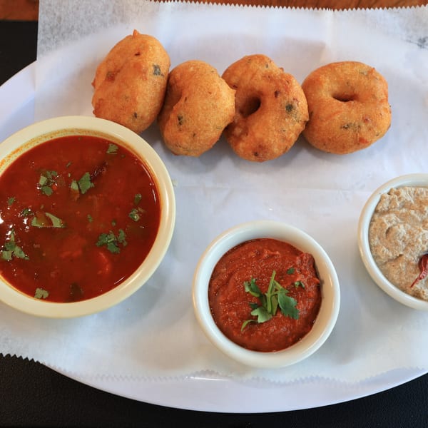 Four golden-brown Indian vada donuts with three bowls of red and beige dipping sauces.