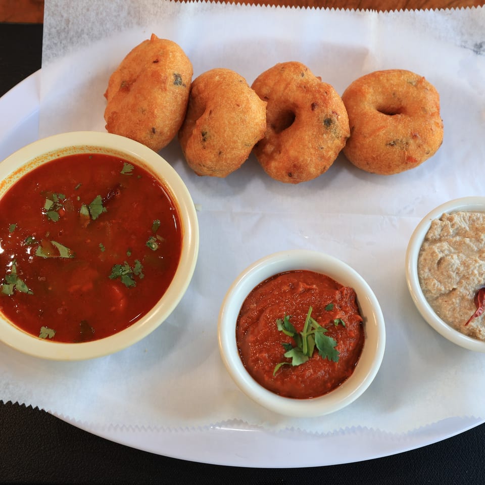Four golden-brown Indian vada donuts with three bowls of red and beige dipping sauces.