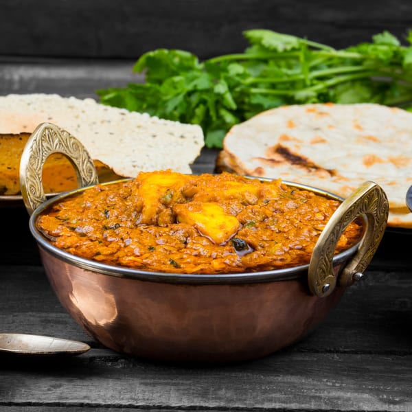Indian paneer curry in a copper bowl, with naan bread and papadums in the background.