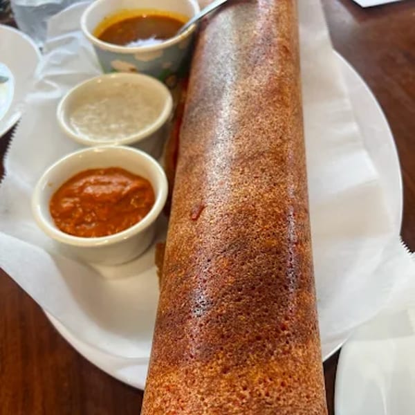 A long, rolled-up dosa with three small bowls of chutneys and sambar on a white plate.